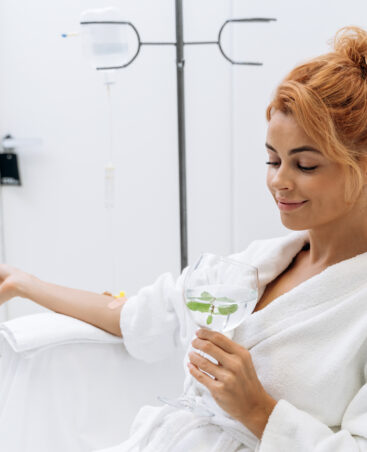 Waist up portrait view of the charming woman in white bathrobe sitting in armchair and receiving IV infusion. She is holding glass of lemon beverage and smiling